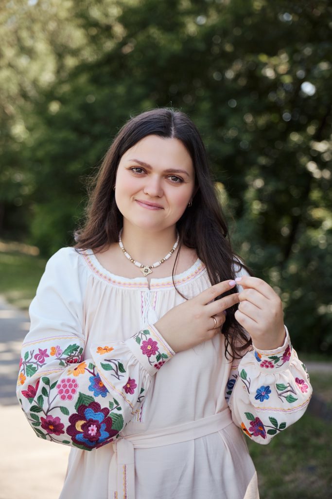 Жінка у світлій сукні з яскравою вишивкою позує на тлі зеленого парку