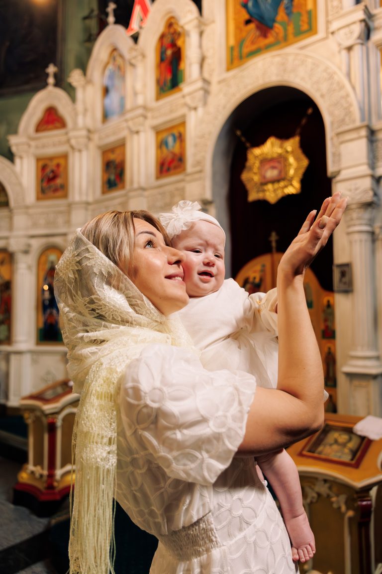 Жінка у білій хустці піднімає усміхнене немовля на тлі іконостаса