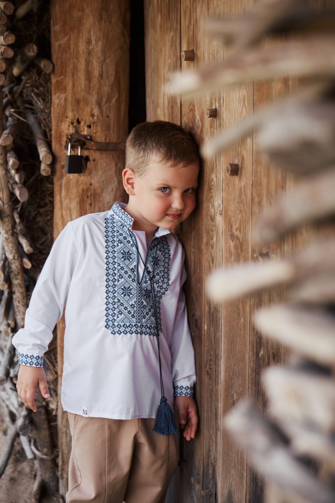 Хлопчик у білій вишиванці стоїть біля дерев'яної стіни старої хати