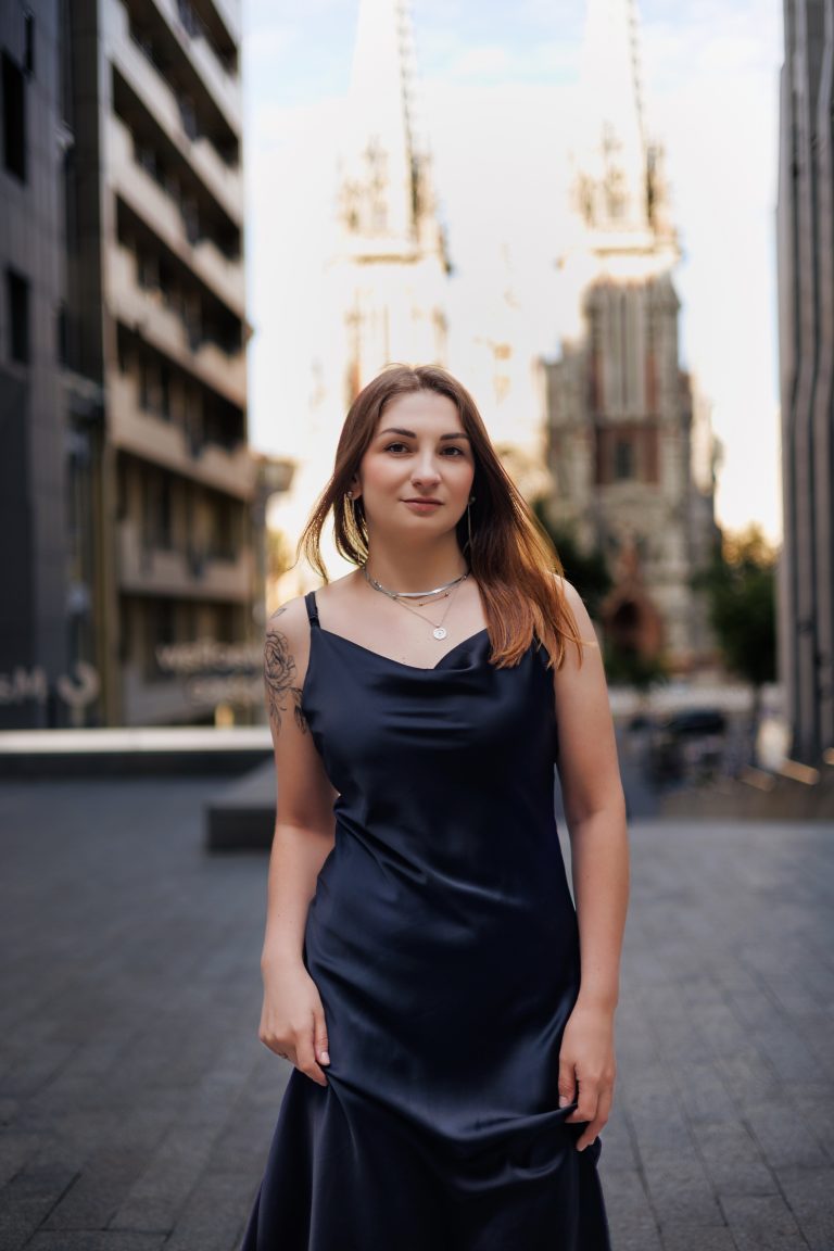 Маргарита у чорній сукні стоїть на міській вулиці на тлі розмитої історичної архітектури