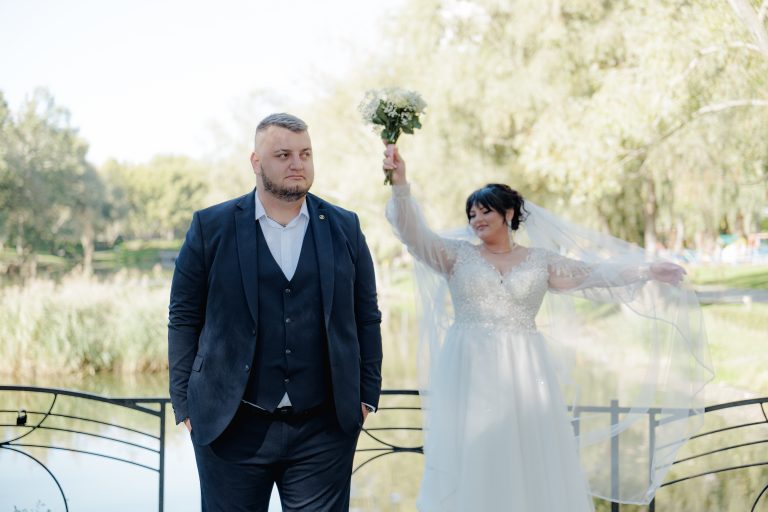 Наречений стоїть біля водойми, а наречена позаду піднімає букет і фату