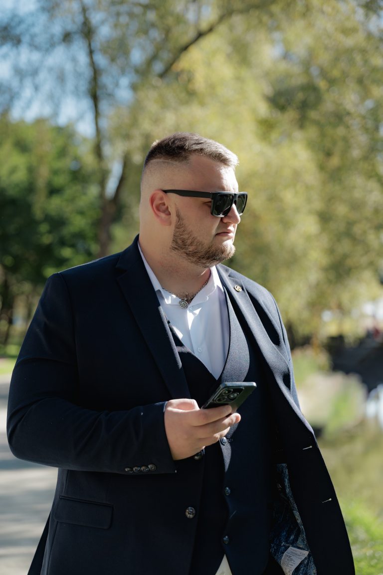 Наречений у темному костюмі та сонцезахисних окулярах стоїть у парку зі смартфоном