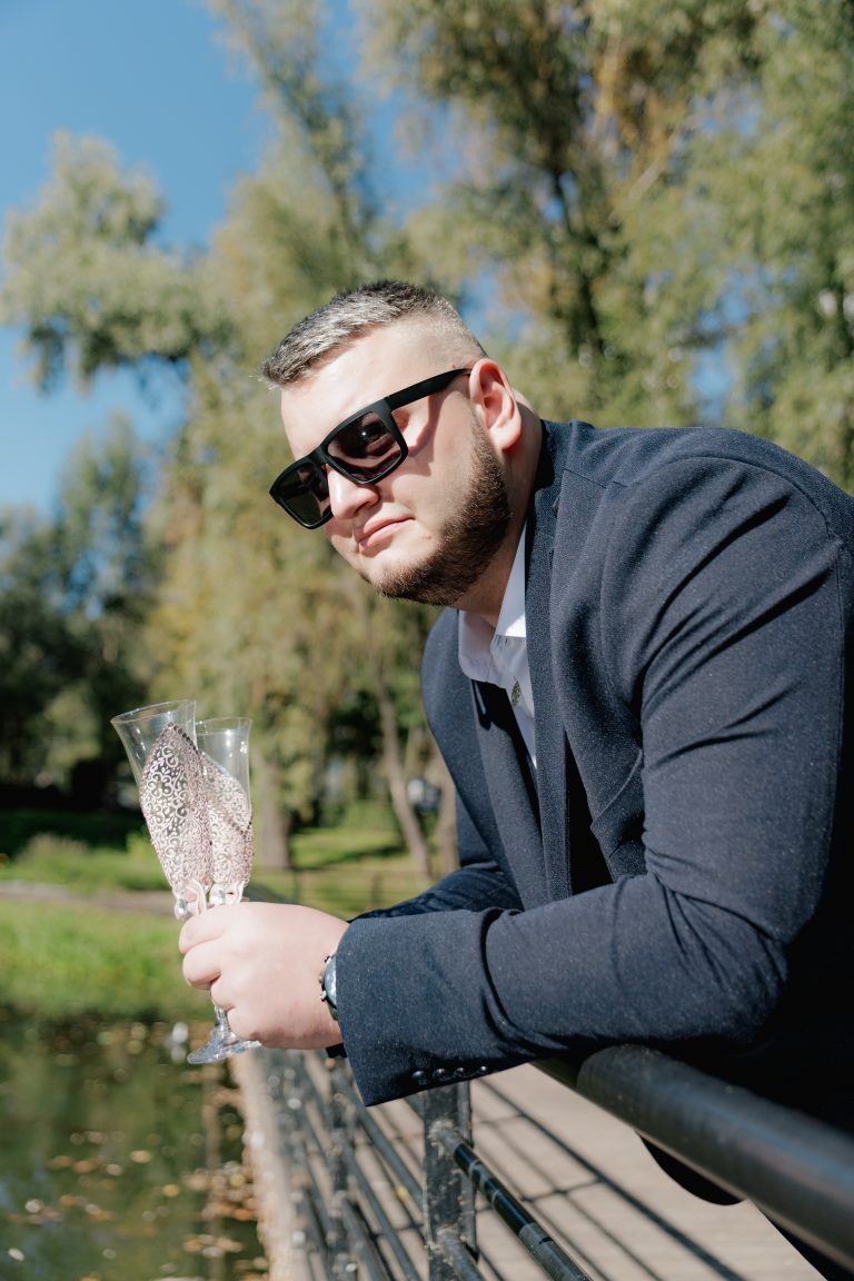 Наречений у сонцезахисних окулярах тримає два декоративні весільні келихи біля водойми