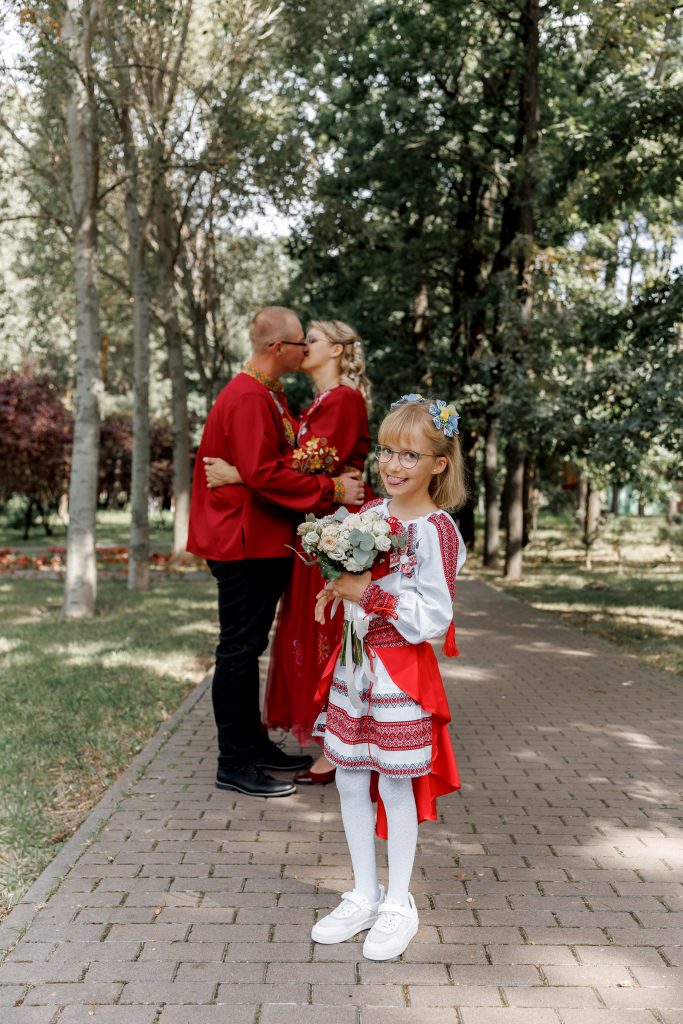 Веронічка у вишиванці стоїть з весільним букетом у парку, а Марина та Ігор цілуються позаду