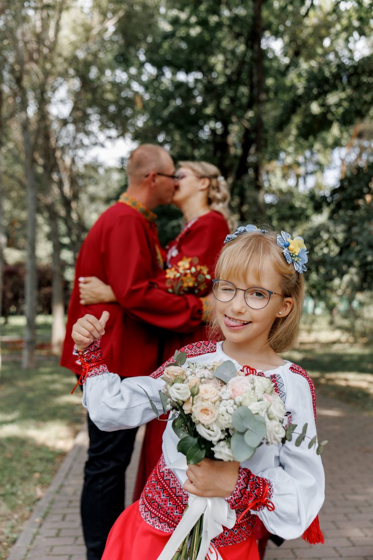 Веронічка з весільним букетом підморгує в камеру, поки Марина та Ігор цілуються на задньому плані