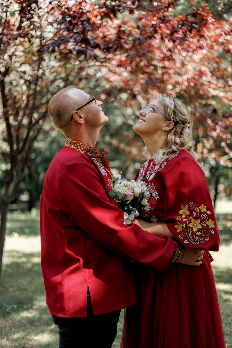 Марина та Ігор у червоних вишитих весільних образах стоять у парку під кроною дерева