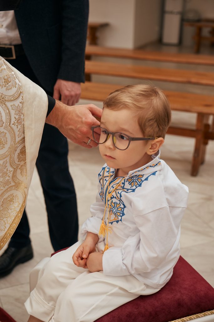Ярослав у окулярах і вишитій сорочці сидить під час церемонії, поруч рука священника