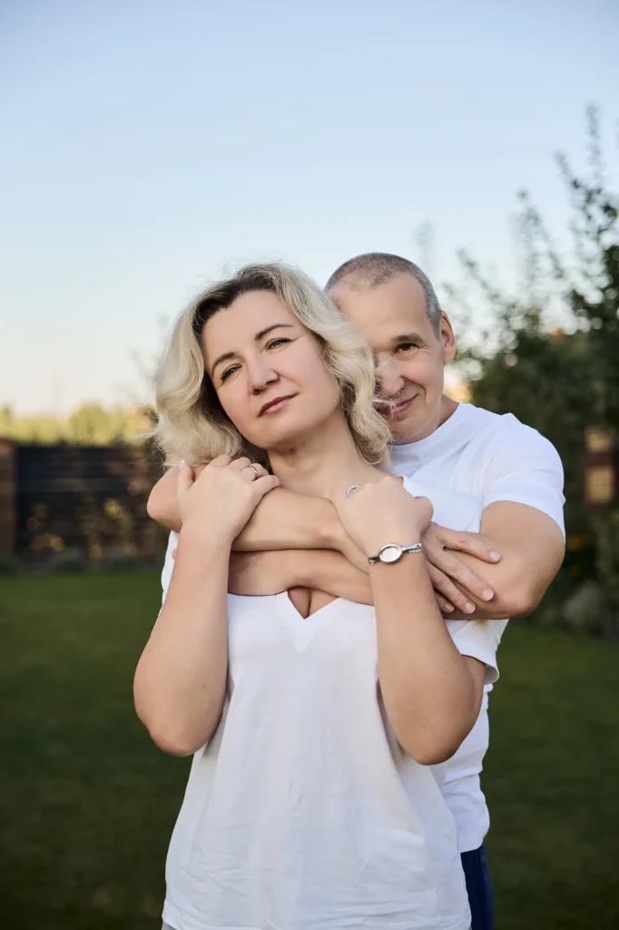 Чоловік обіймає ззаду світловолосу жінку в білій футболці, обоє дивляться в камеру, Київ