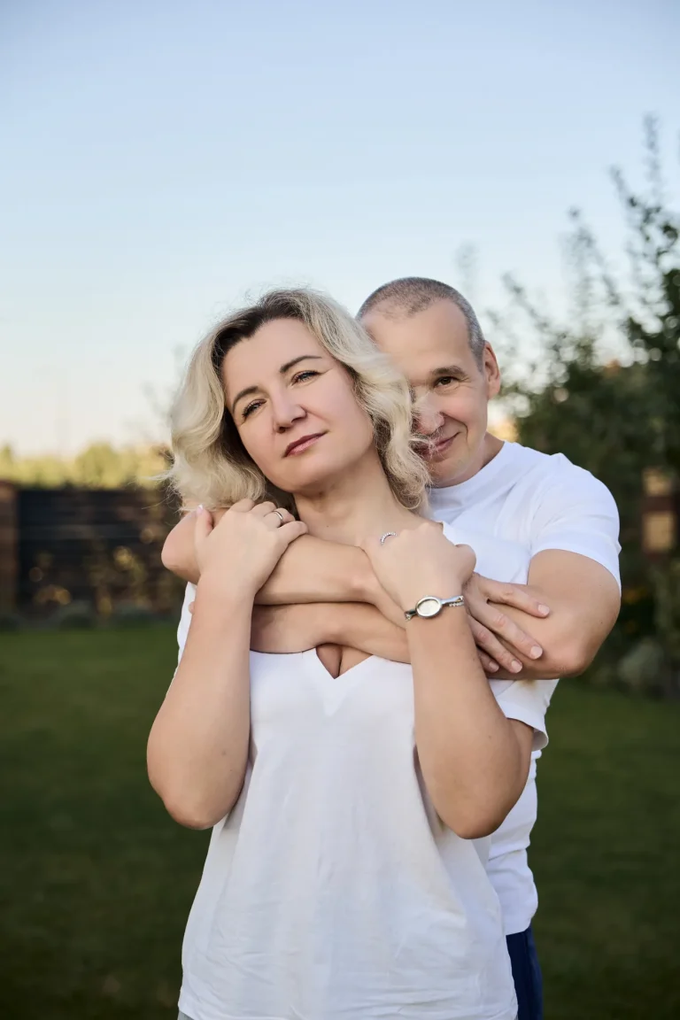 Чоловік обіймає ззаду світловолосу жінку в білій футболці, обоє дивляться в камеру, Київ